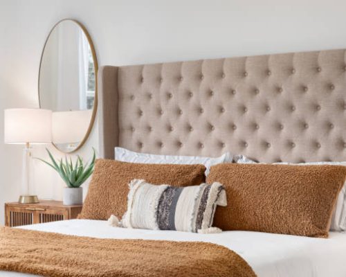 A bedroom detail shot with a cushioned headboard, a gold mirror on the wall, and a plant and lamp on the nightstand.